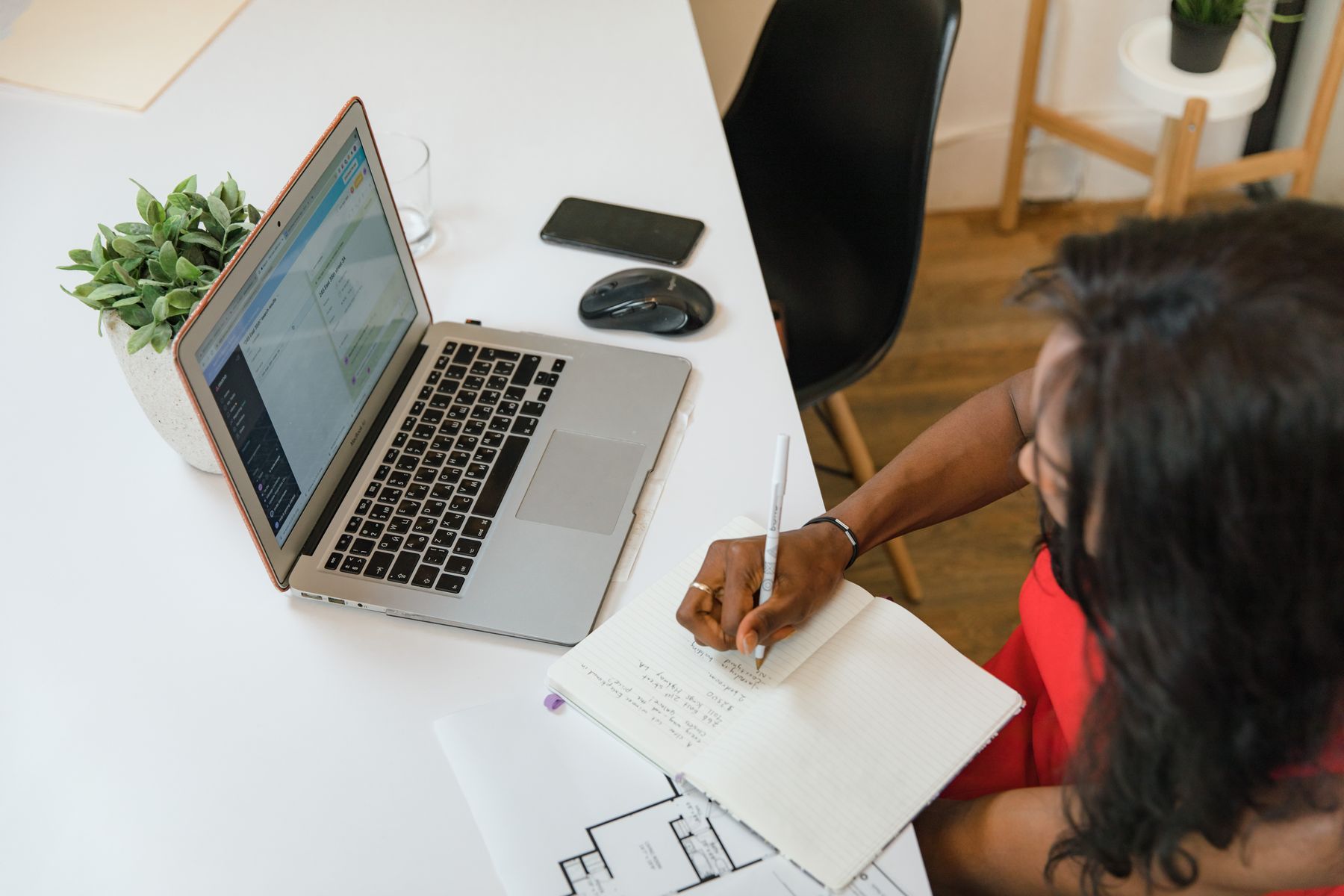 A Black woman business owner writes in a notebook beside her open laptop at a white desk.